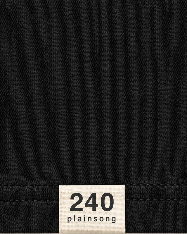 Close-up of the front hem on the black tee: a cream woven label sewn into the hem seam, with 240 in large figures and plainsong in small letters.